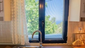 A kitchen with a stainless steel sink, a window with a view of greenery, and a door with a dark frame. - Irene's Cottage Myrtonas (Karpathos)