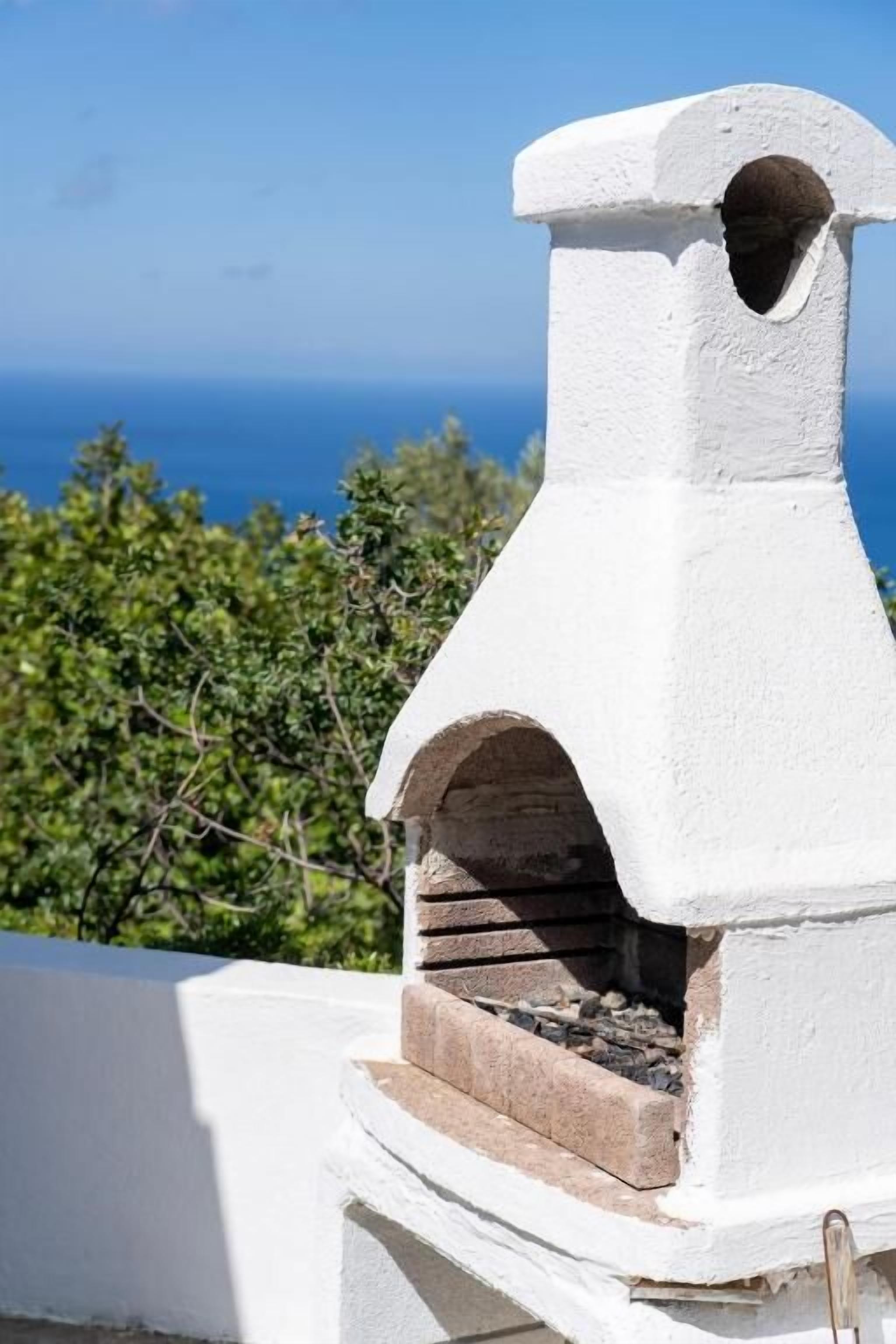 Un horno de piedra blanco con base de ladrillo, frente a un fondo de vegetación y el mar.