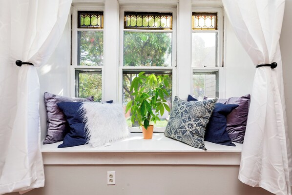 Ein Fensterplatz mit Blumentopf, gemusterten Kissen und einem weißen Vorhang.