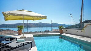 A pool with lounge chairs and an umbrella, overlooking a body of water and hills. - Luxury apartment with stunning views and pool (Seget Vranjica)