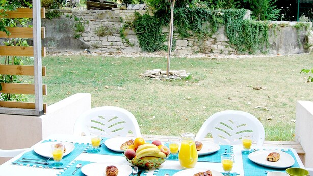 Un área de comedor al aire libre con una mesa preparada para el desayuno, que incluye platos con comida, un frutero y una jarrón de jugo de naranja.