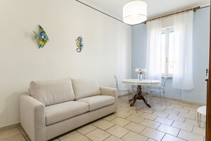 Ein Wohnzimmer mit einem beigen Sofa, einem kleinen runden Tisch mit Stühlen, einem Fischbild an der Wand und einem Fenster mit durchscheinenden Vorhängen.