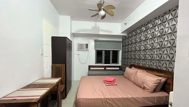 Ein Schlafzimmer mit einem Bett, einem Schreibtisch, einem Stuhl, einem Kleiderschrank, einem Deckenventilator, einem Fenster und einer Wand mit geometrischem Muster.