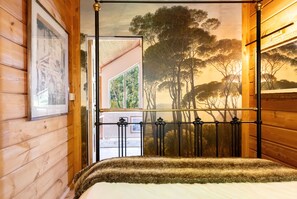 Ein Schlafzimmer mit einem Bett, einer Holzwand, einem Gemälde und Blick auf Bäume durch ein Fenster.