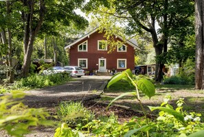 Uma casa vermelha com varanda frontal, rodeada por árvores e um caminho de acesso de cascalho.