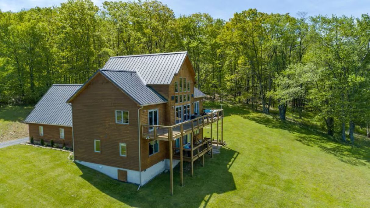 A two-story wooden house with a balcony, surrounded by a grassy area and trees.
