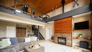 En rummelig opholdsstue med højt til loftet, pejs, trappe og et tv ophængt på væggen.
