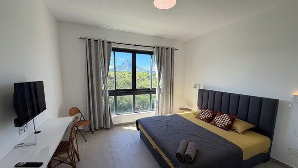 A hotel room with a bed, a desk, a TV, and a window with curtains. - J'Vais Residence Flic en Flac (Flic en Flac)
