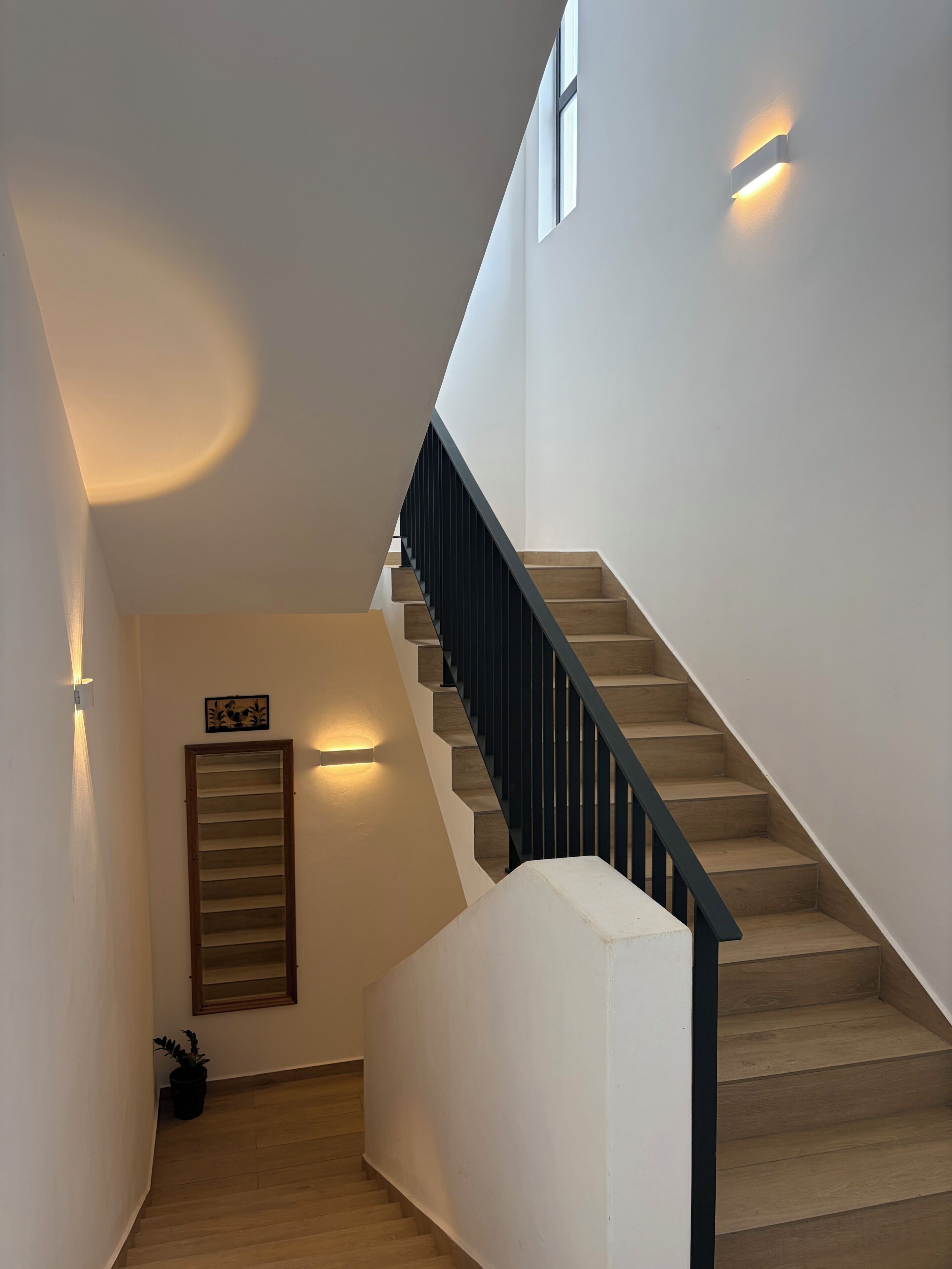 Eine moderne Treppe mit Holzhäuschen und einem schwarzen Geländer, die zu einer Zwischenebene mit einer Leuchte und einem gerahmten Bild führt.