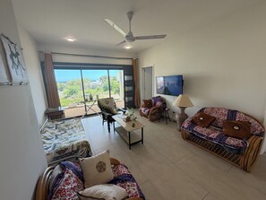 A spacious living room with a large window, a ceiling fan, a television, and a sofa with patterned cushions. - J'Vais Residence Flic en Flac (Flic en Flac)