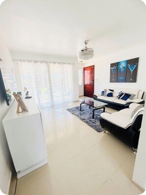 A modern living room with a sofa, a coffee table, a chandelier, and a door. - “ Enjoy Your Coffee with Poolside Views at Harmony II – Balcony Bliss" (Santiago de los Caballeros)