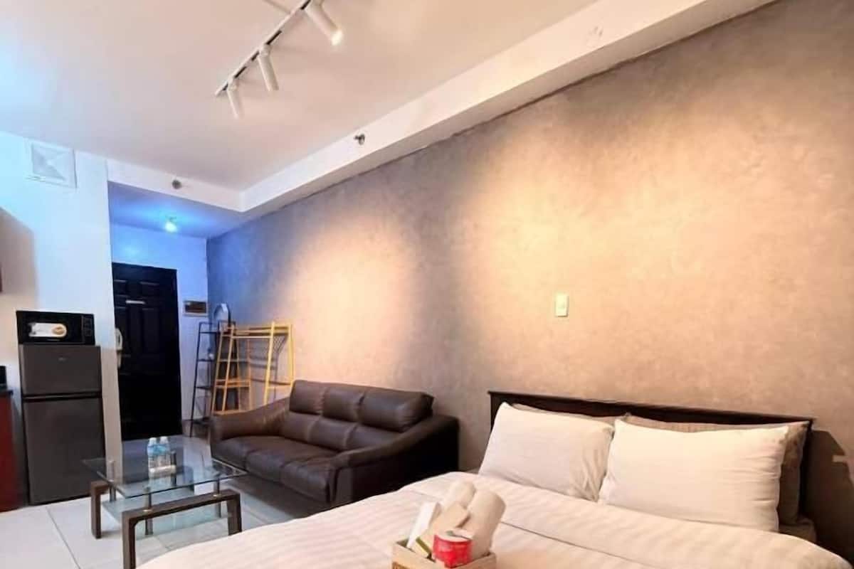 A bedroom with a large bed, a glass coffee table, a sofa, and a kitchenette in the background.