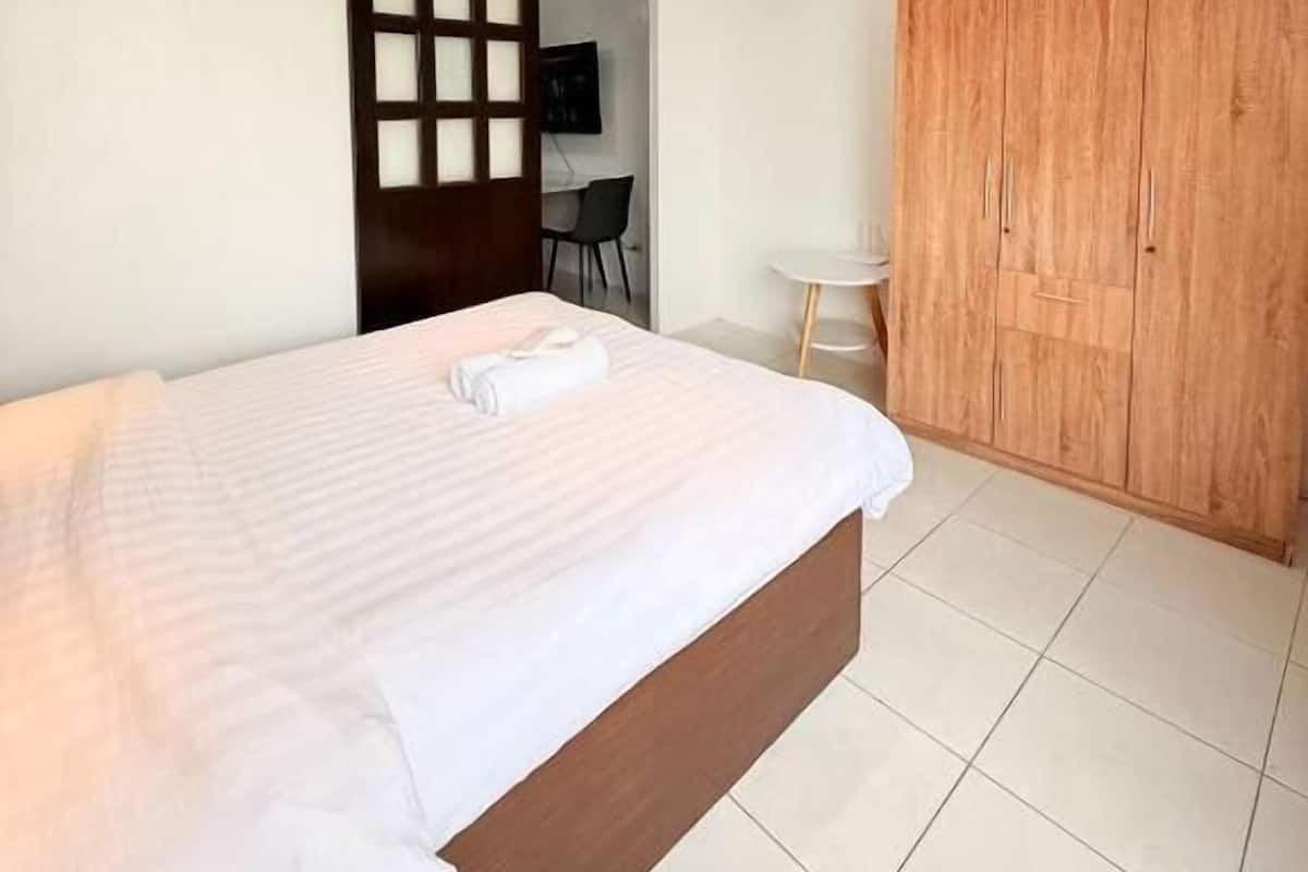 A bedroom with a bed, a wooden wardrobe, a small table, and a door leading to another room.