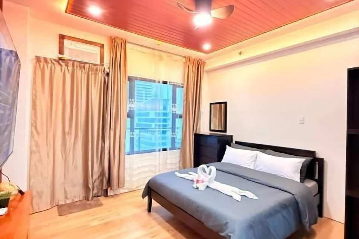 A hotel room with a bed, wooden flooring, a ceiling fan, and a window with curtains.