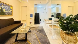 Una sala de estar moderna con una mesa de centro de vidrio, un sofá, un cuadro grande, una planta en maceta y una cocina al fondo.