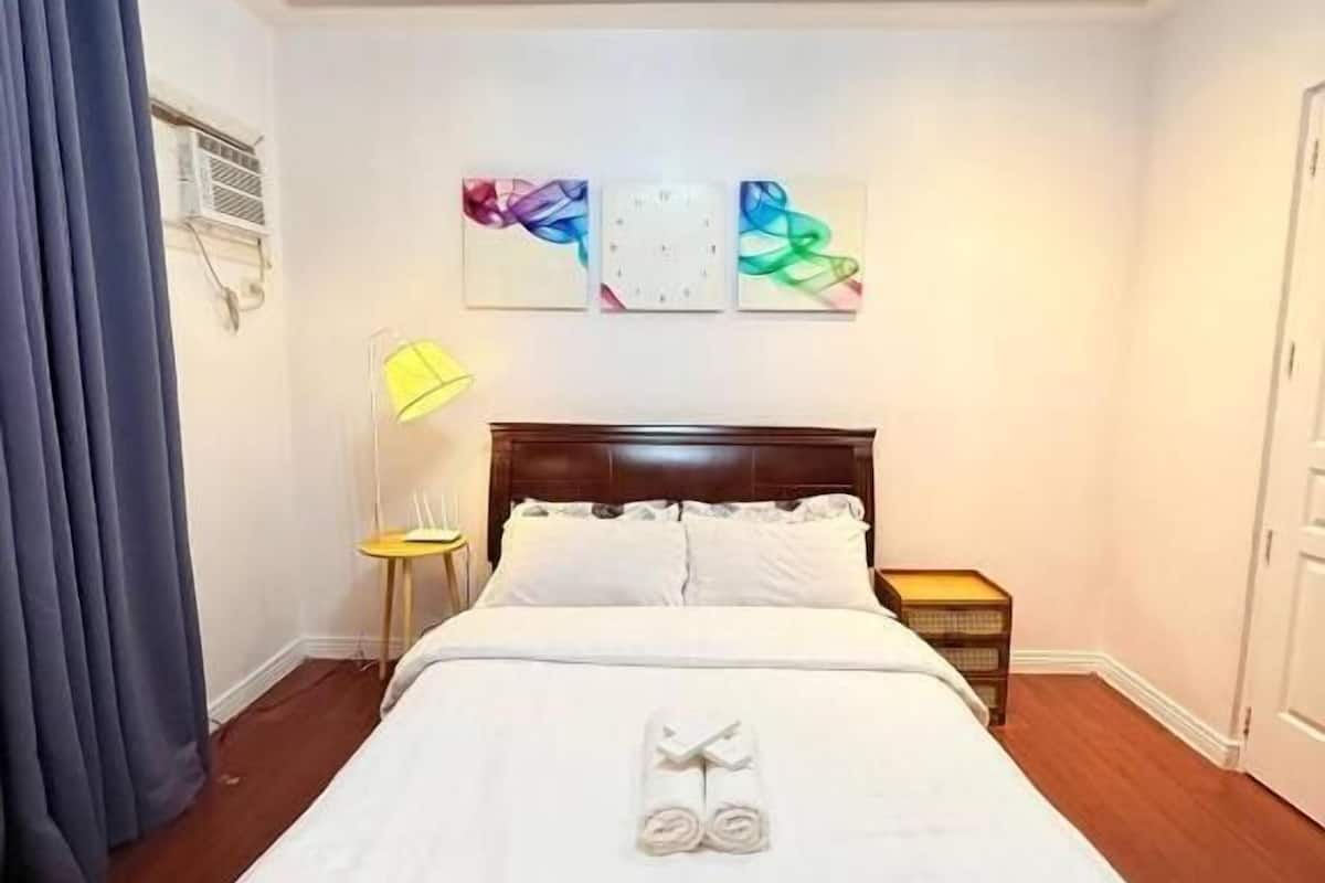 A hotel room with a bed, a bedside table, a lamp, a wall clock, and a door.