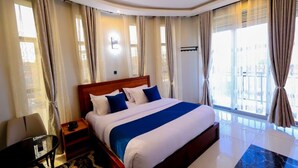 A hotel room with a large bed, a nightstand, a chair, and a view of the city through large windows. - Sea bridge view hotel mbarara (Mbarara)