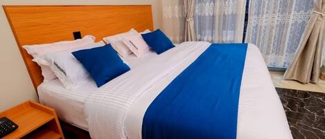 Ein Hotelzimmer mit hölzernem Kopfteil, einem Bett mit weißer und blauer Bettwäsche, einem Nachttisch mit Fernbedienung und Blick durch transparente Vorhänge.