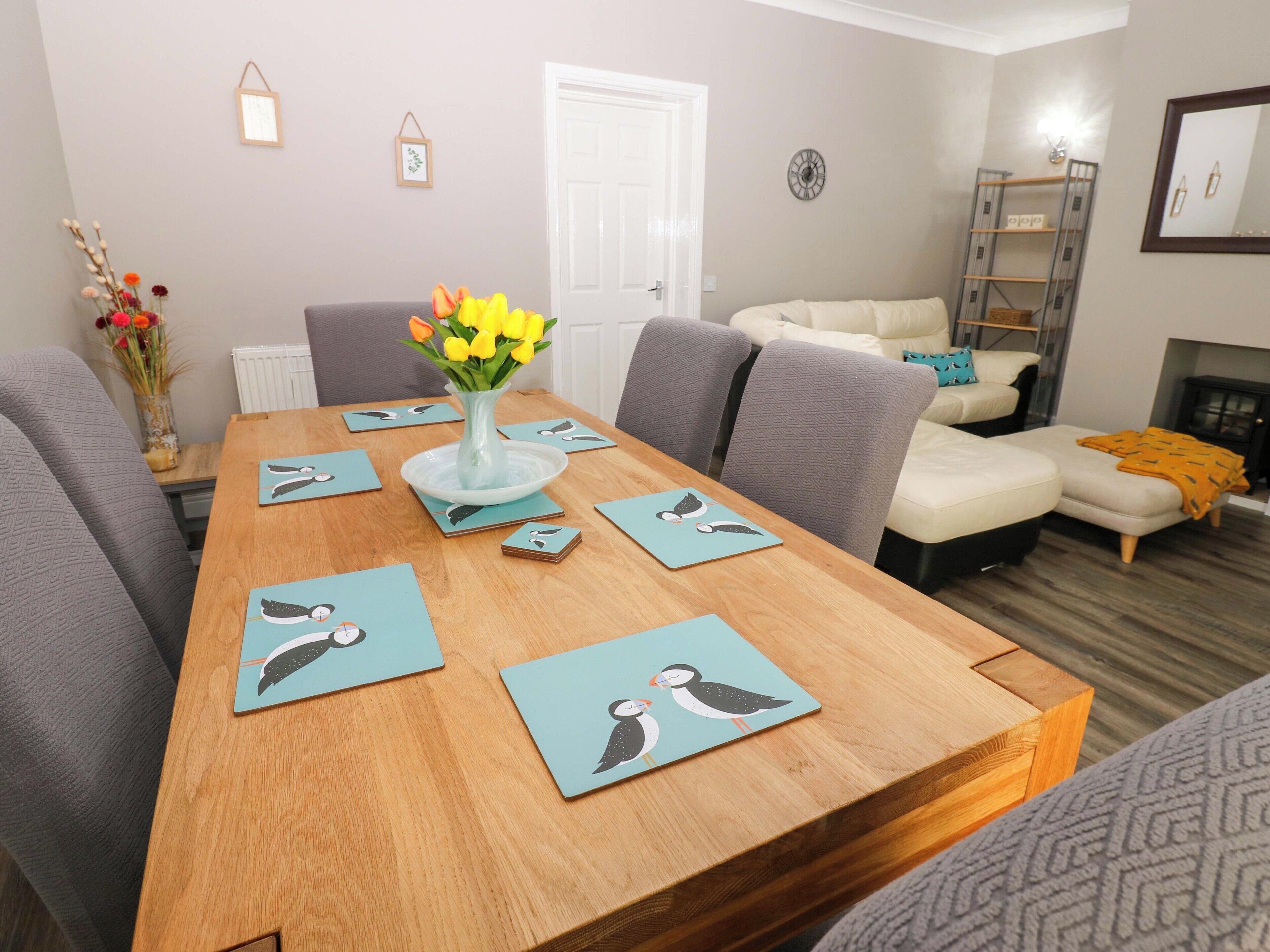 A dining area with a wooden table, grey upholstered chairs, and a vase with yellow flowers.