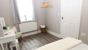 Un dormitorio con cama, escritorio y silla, radiador, ventana con cortinas, puerta y perchero.