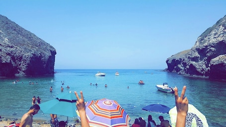 A beach with people swimming and boats in the water, surrounded by rocky cliffs.. complexe touristique village des fourmis