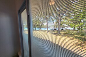 Ein Fenster mit horizontalen Rollläden, Blick auf einen Parkplatz mit Bäumen und einer Bank, sowie ein Gebäudefassadenbild.