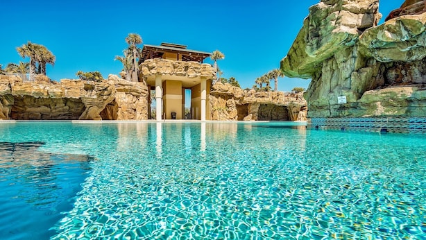 Una piscina amb aigua cristal·lina, un edifici amb façana de pedra i palmeres al fons.