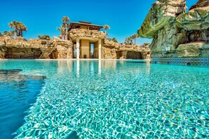 A pool with clear water, a building with a rock facade, and palm trees in the background. - ☀️Golf Cart | Resort Pool | 30A | Gulf Breeze☀️ (Santa Rosa Beach)