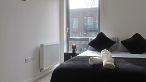 Un dormitorio con un ventanal grande, un radiador, una mesita de noche con una lámpara y una cama bien hecha con ropa de cama blanco y negro.