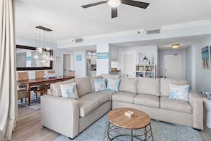 A living room with a sectional sofa, a dining table with chairs, a ceiling fan, and a kitchen area in the background. - ⭐Across the Street from the Beach-Great, Spacious Layout-2BR Enclave 701A⭐ (Destin)