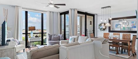 Amplia sala de estar con sofá, mesa de comedor y sillas, ventanas grandes con cortinas y ventilador de techo.