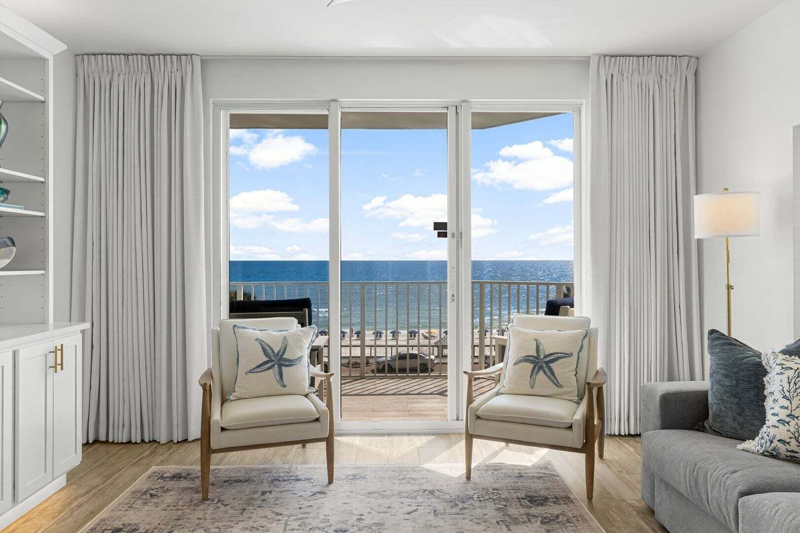 A room with a view of the ocean, a balcony with outdoor furniture, and a sofa with decorative pillows.