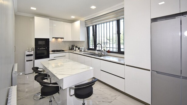 Una cocina moderna con armarios blancos, encimera de mármol, horno empotrado, refrigerador de acero inoxidable y una ventana que proporciona luz natural.