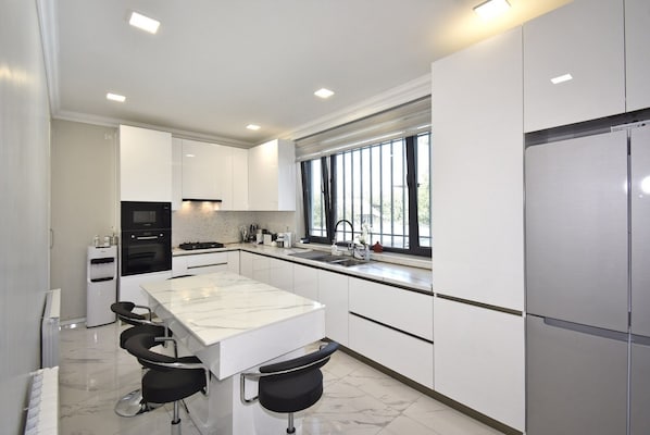 A modern kitchen with white cabinetry, a marble countertop, a built-in oven, a stainless steel refrigerator, and a window providing natural light.