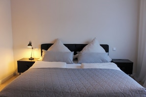 A neatly made bed with a quilted cover, two bedside tables with lamps, and a plain wall.