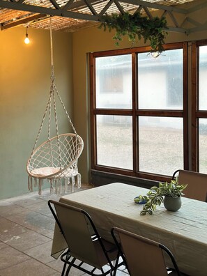 A room with a hanging chair, a table with chairs, a potted plant, and a large window.