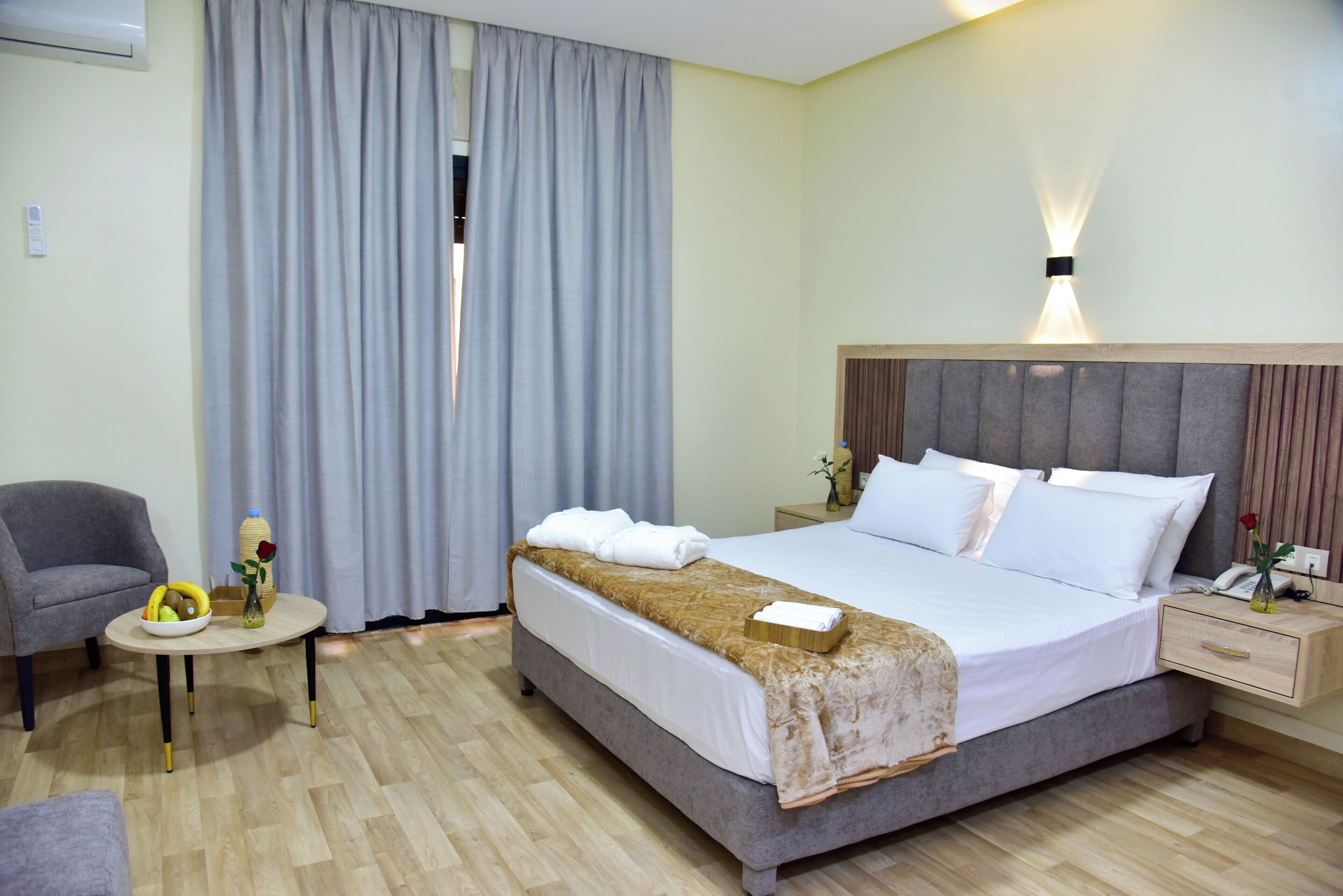 A hotel room with a bed, a chair, a small table with a fruit bowl and vase, and a nightstand with a phone and vase.