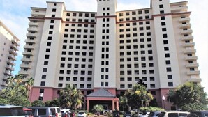 A high-rise hotel with multiple floors, a central entrance, and a parking lot with several cars. - Beach Club D209- Direct Gulf View - Beach - Pool - WIFI (Gulf Shores)
