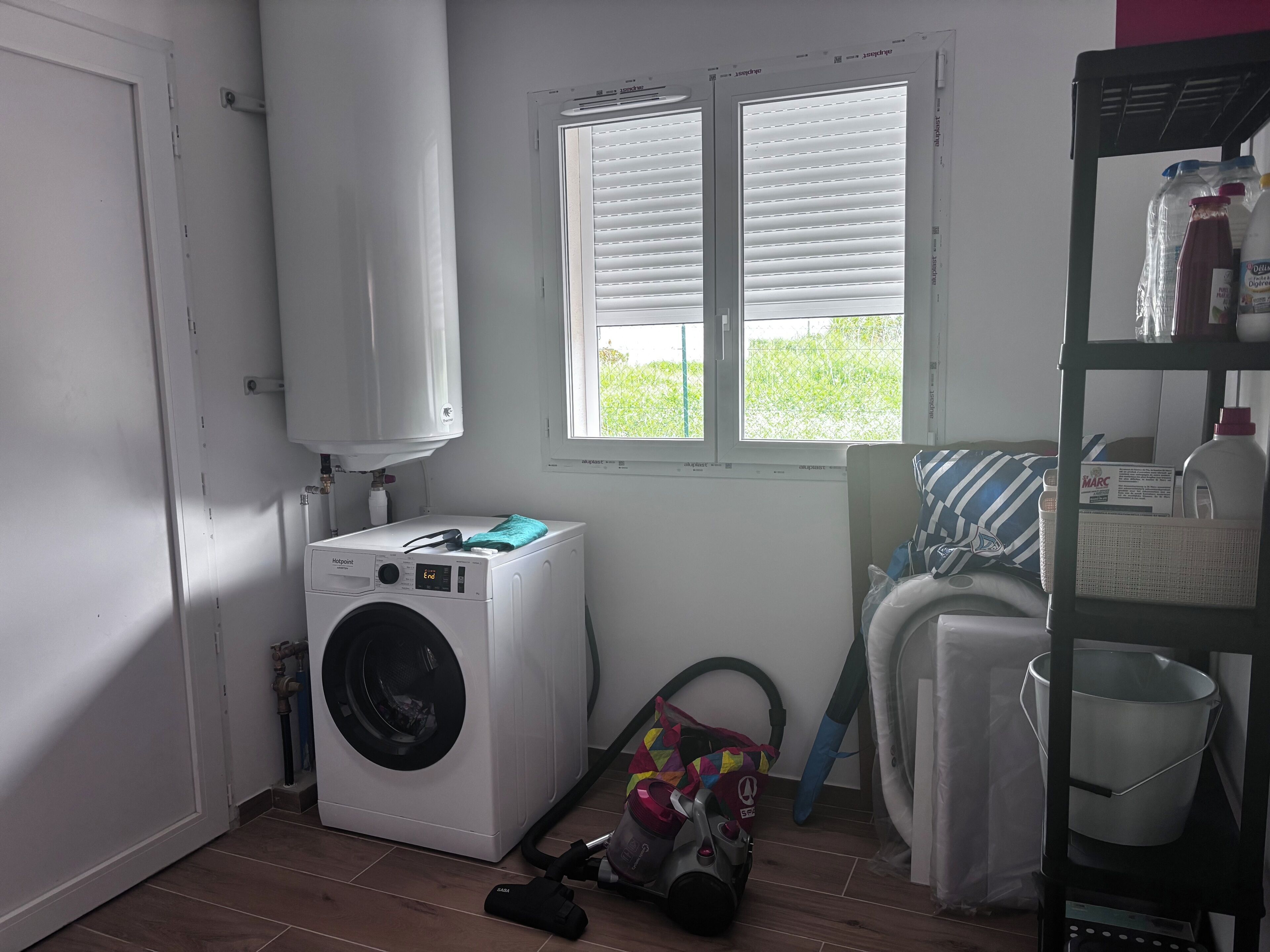 A laundry room with a washing machine, a window with blinds, a vacuum cleaner, and a shelf with bottles.