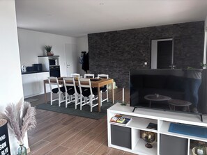 A modern living room with a TV, dining table, chairs, and a kitchen area in the background. - House with large garden for 4/6 people (Aléria)