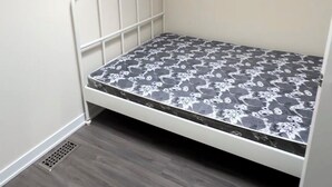 Habitación pequeña con una cama individual de estructura metálica, colchón estampado, suelo de madera y lámpara colgante.