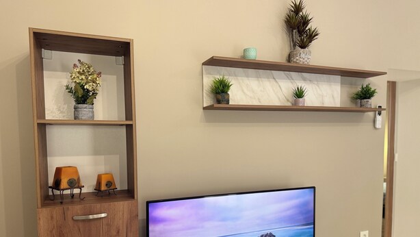 A television on a wooden stand with a remote, a small round table, and two shelves with decorative items. - Modern cozy home in Kos town (Kos)