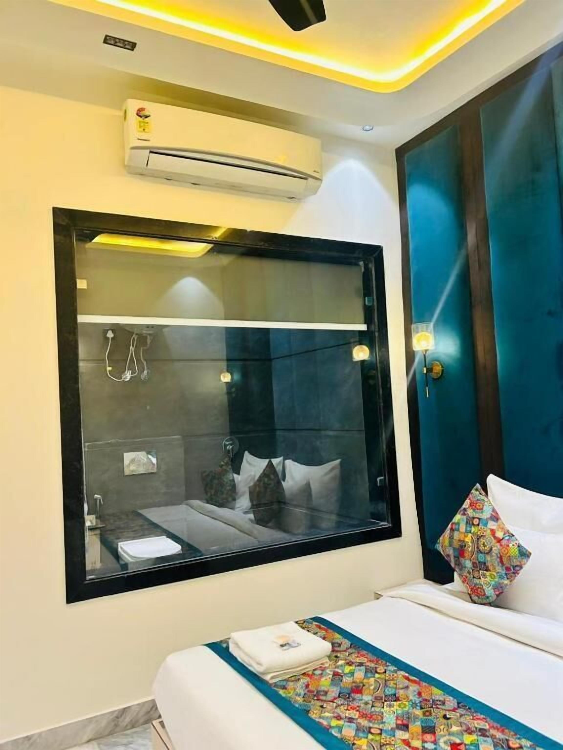 A modern hotel room with a large window, a bed with white linens, a colorful quilt, and an air conditioning unit on the wall.