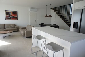 Une cuisine moderne dotée d’un îlot central, de tabourets de bar, d’un coin repas avec une table et des chaises, d’un escalier et d’un salon avec un canapé et une table basse.