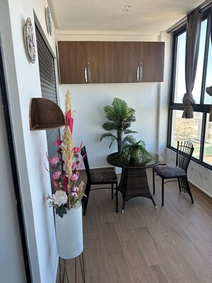 Ein Zimmer mit Esstisch, Stühlen, einer Topfpflanze, einem Wandregal und einem Fenster mit Vorhängen.