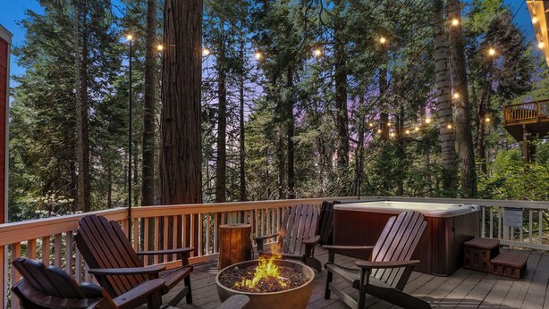 Eine Terrasse mit Adirondack-Stühlen, einem Feuerkorb und einem Whirlpool, umgeben von hohen Bäumen.