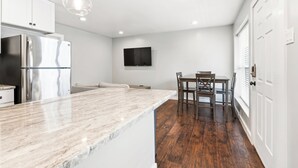 A modern kitchen with a marble countertop, stainless steel appliances, a dining area with wooden chairs, and a flat-screen TV mounted on the wall. - Glendale Court Unit 6 (Dallas)