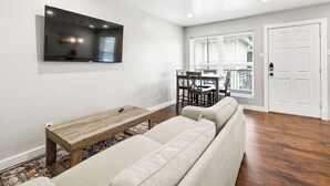 A living room with a flat-screen TV, a wooden coffee table, a sofa, a dining table with chairs, and a door. - Glendale Court Unit 6 (Dallas)