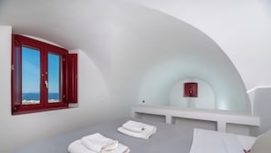 A bedroom with a bed, two towels, a window with red shutters, and a view of the sea. - Zen Villas Oia (Oia)