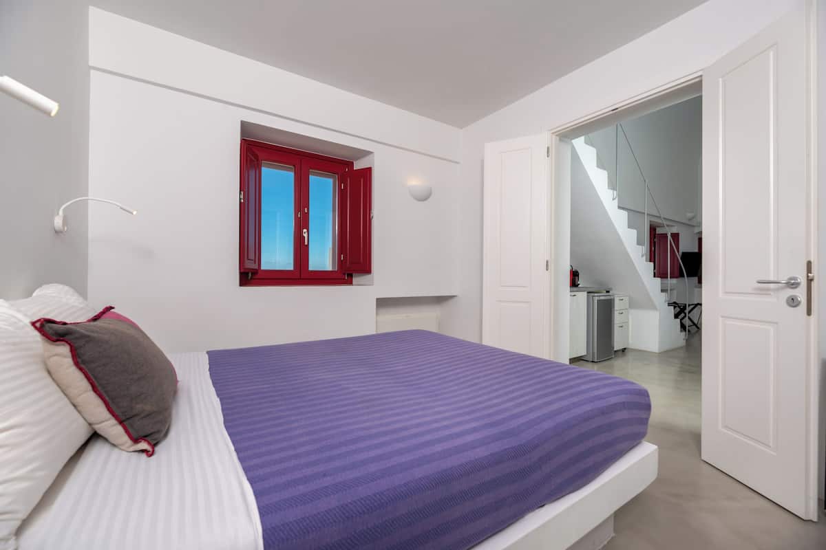 Une chambre à coucher avec un lit, une fenêtre avec des volets rouges, une porte blanche et un escalier menant à un espace cuisine.